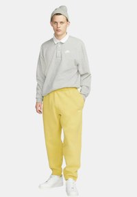 Grå långärmad polo med vit krage och Nike-logotyp, ihop med ljusgula träningsoverallsbyxor och vita sneakers, bärs med en grå mössa.