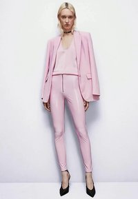 Femme se tenant contre un mur blanc, portant un pantalon rose brillant, un blazer rose assorti et un haut, un collier ras du cou noir et des talons noirs.