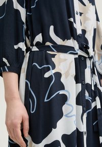 Blumenmuster-Kleid in Marineblau, Weiß und Beige. Hergestellt aus einem glatten Stoff, hat es eine Knopfleiste vorne und einen passenden Gürtel in der Taille.