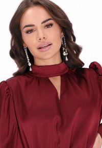 Abito di raso bordeaux con scollatura alta e taglio a chiave. Maniche a sbuffo e una texture liscia. Orecchini pendenti con perle e cristalli.