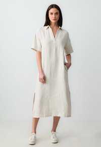 Linnen jurk in een lichtbeige kleur, met korte mouwen, een kraag, zakken en zijsplitten. Gekozen in combinatie met witte sneakers.