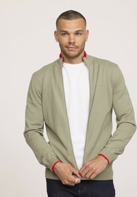 Veste zippée vert clair avec un col intérieur rouge, des poignets et un ourlet côtelés ; portée sur un t-shirt blanc. Présente un petit logo sur la poitrine.