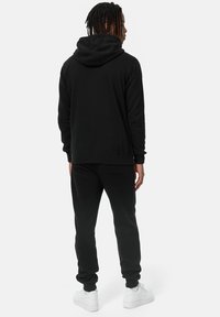 Sættet består af en sort hættetrøje og sweatpants lavet af blødt fleece-materiale. Det har en stor hætte, ribbede manchetter og en afslappet pasform. Bæres med hvide sneakers.