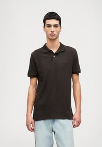 Καφέ πόλο μπλουζάκι με επίπεδο γιακά και κοντούς μανικούς. Διαθέτει τριπλό κουμπί και διακριτό κεντημένο λογότυπο στο στήθος. Χαλαρή εφαρμογή.