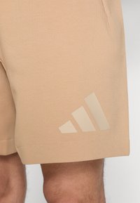 Peach-colored athletic shorts made of soft fabric, featuring a prominent light beige Adidas logo on the left side and side pockets.
