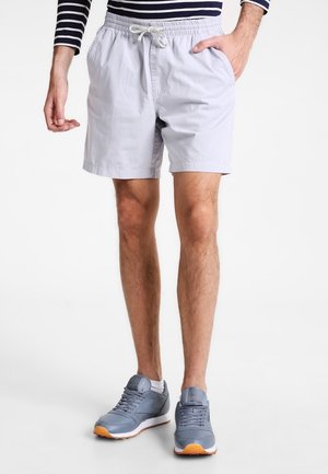 Mann trägt hellgraue Shorts mit Kordelzug, ein navy-blau gestreiftes Langarmshirt und blaue Sportschuhe und steht vor einem weißen Hintergrund.