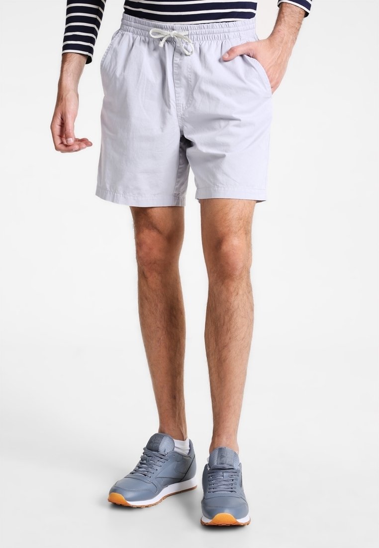 Homme portant un short en tissu léger gris clair, une chemise à manches longues rayée navy, et des chaussures de sport bleues se tenant sur un fond blanc.