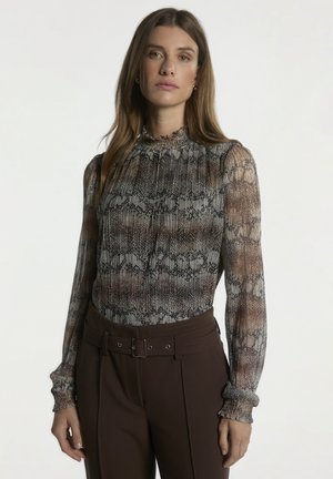 Vrouw draagt een blouse met lange mouwen, hoge hals en slangenprint en een donkerbruine broek met hoge taille en riem, staand tegen een effen achtergrond.