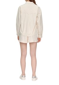 Cremefarbene, gestreifte Bluse mit langen Ärmeln, Rückenpasse-Design, kombiniert mit passenden Shorts und weißen Turnschuhen mit transparenten Fersenakzenten.