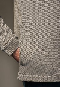 Personne portant un vêtement à manches longues texturé beige et noir, la main glissée dans la poche latérale, sur un fond neutre uni.