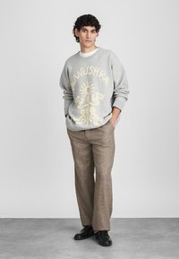 Siv svidar s puloverjem v zlatem grafičnem oblikovanju in besedilu, v kombinaciji s sivimi strukturiranimi hlačami in črnimi čevlji. Model stoji proti nevtralni ozadju.