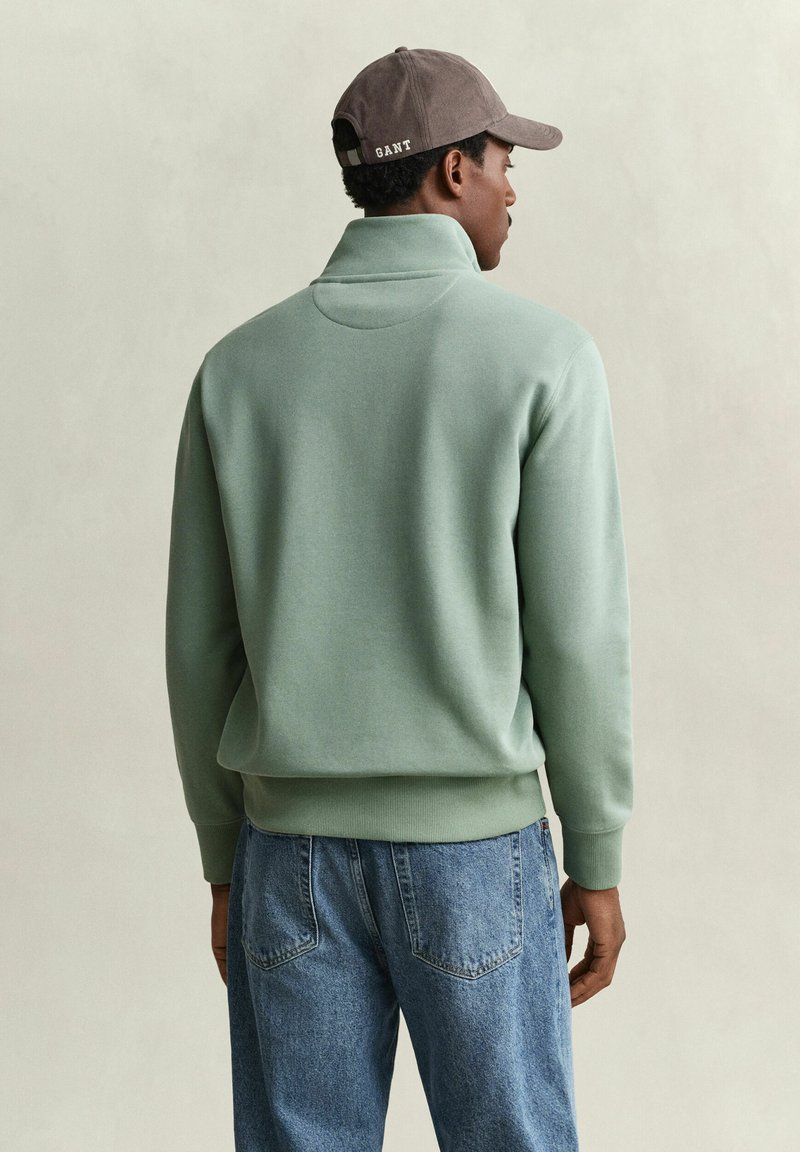 Man wearing a light green pullover, blue jeans, and a gray cap with "GANT" text, shown from behind against a plain background.
