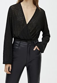 Blouse noire à manches longues en tissu transparent et texturé. Associée à un pantalon en simili cuir noir à taille haute et brillant, avec des poches latérales.
