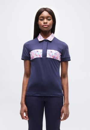 Žena v tmavomodrej košeli s krátkymi rukávmi a farebnými vzorovanými golierom a panelmi na hrudi, stojaca pred jednoduchým svetlým pozadím.