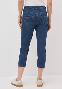 Pantalons capri en denim bleu foncé avec une silhouette ajustée, deux poches arrière et des coutures visibles. Associés à des baskets blanches.