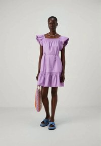 Vestido roxo com mangas com babados, cintura franzida e saia em camadas. Combinado com sandálias azuis e uma bolsa de rede multicolorida.