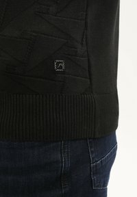 Juodas tekstūruotas megztinis su geometriniais raštais virš siauro apdailos, dėvimas virš tamsiai mėlynų džinsų su matomu užpakaliniu kišeniu.