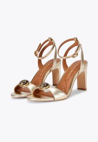 Złote metaliczne sandały na wysokim obcasie z otwartym noskiem, pasek wokół kostki i ozdobnymi okrągłymi akcentami z przodu. Płaska podeszwa, teksturowane wnętrze.