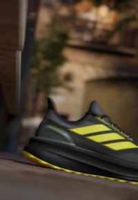 Baskets noires à texture lisse, avec des accents jaune vif et trois bandes sur le côté. Semelle en caoutchouc offrant une bonne adhérence.