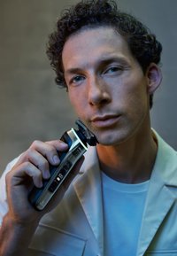 Elektrische scheermes met een metallic zilveren en donkerblauwe afwerking. Het digitale display toont een batterijniveau van 100%. Gebogen ontwerp en een zwarte scheerkop.