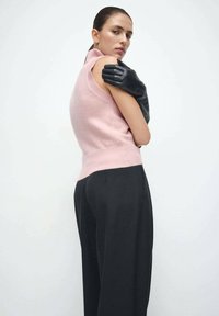 Svijetloružičasti pleteni top bez rukava s visokim ovratnikom upareni s crnim hlačama visokog struka. Na ramenu počiva crna rukavica.