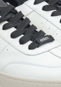 Witte leren sneakers met zwarte veters en een zwart label met de tekst "estro." De zool is beige met een gestructureerde afwerking.