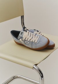 Baskets bleu clair texturées avec lacets blancs et semelles marron placées côte à côte sur une chaise en cuir beige avec cadre métallique.