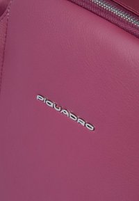 Rosa Ledertasche mit einem glänzenden silbernen "Piquadro"-Logo und einer strukturierten Oberfläche, oben mit Reißverschluss.