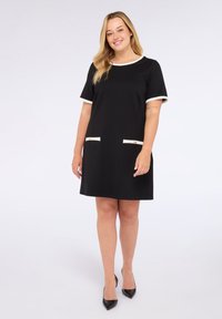 Vestido negro con mangas cortas y ribete blanco en el escote y los puños. Presenta dos bolsillos blancos con botones dorados.