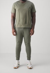 Bavlněné tričko olivově zelené barvy s výstřihem do V a malým logem, doplněné o odpovídající zúžené tepláky. Obuv má bílý a zelený design.