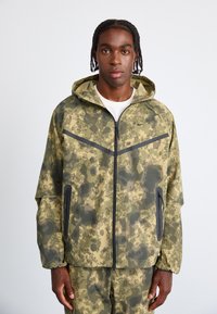 Camouflagemønsteret jakke i nuancer af olivengrøn og sort, med lynlåslukning, hætte og sidelommer med kontrastkant.