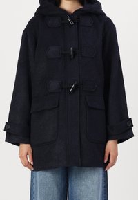 Abrigo tipo duffle con capucha en azul marino, con cierres tipo toggle, bolsillos con solapa y tirantes en los puños con botones, llevado sobre unos pantalones de mezclilla azules.