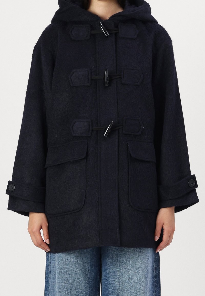 Abrigo tipo duffle con capucha en azul marino, con cierres tipo toggle, bolsillos con solapa y tirantes en los puños con botones, llevado sobre unos pantalones de mezclilla azules.