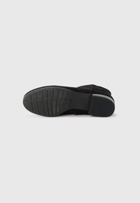 Schwarze Wildlederschuhe mit einer strukturierten Gummisohle, die eine abgerundete Spitze und einen niedrigen Absatz aufweisen. Das Innere ist weich und gepolstert für zusätzlichen Komfort.
