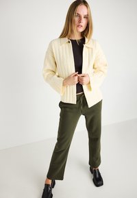 Kremowy sweter w warkocz z kołnierzem, połączony z oliwkowo-zielonymi spodniami dresowymi i czarnymi teksturowanymi butami. Prosty, codzienny styl.