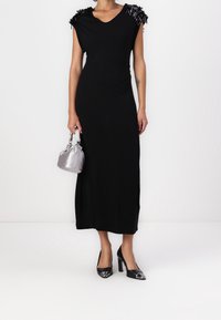Robe maxi noire à coupe ajustée, décolleté en V et accents de franges texturées sur les épaules. Associée à un sac à main argenté et des chaussures à talons hauts noires.