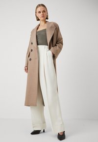 Cappotto doppiopetto marrone chiaro con grandi bottoni, indossato su un top verde scuro e pantaloni plissettati color crema, abbinato a scarpe col tacco alto nere.
