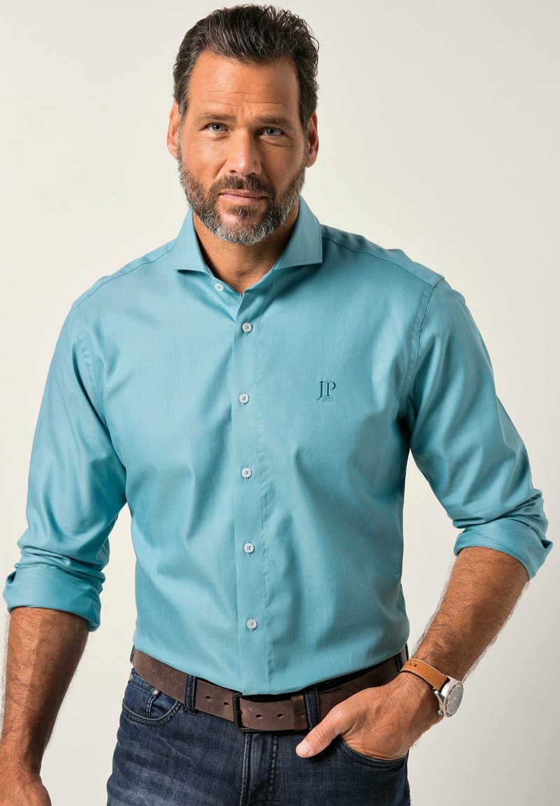 Muž s plnovousem, oblečený v teal košili s krátkým rukávem, s hnědým páskem, džíny a hodinkami, s rukou v kapse, na pozadí bez vzoru.