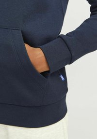 Temno modra majica s kapuco z mehko teksturo, ki ima kenguru žep in rebrast rokav. Majhna oznaka logotipa je vidna blizu šiva žepa.