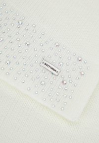 Tissu blanc tricoté avec un motif texturé, orné de multiples clous irisés et d'une étiquette en argent portant l'inscription "WITTCHEN".