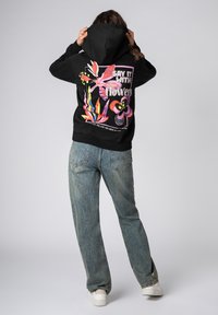 Zwarte hoodie met een kleurrijke bloemenafbeelding op de achterkant en tekst. Gecombineerd met lichtblauwe wijde jeans en witte sneakers.