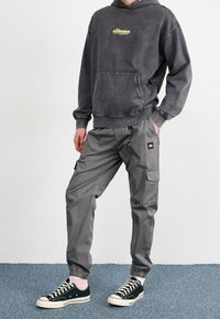 Person som bär en utsliten svart ellesse-hoodie, grå cargobyxor med logotypmärke, vita strumpor och svarta låga sneakers står på blå matta.