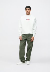 Bílá mikina s červeným a modrým logem "TOMMY", doplněná olivově zelenými kalhotami širokých nohavic a bílými teniskami; model stojí před jednoduchým pozadím.