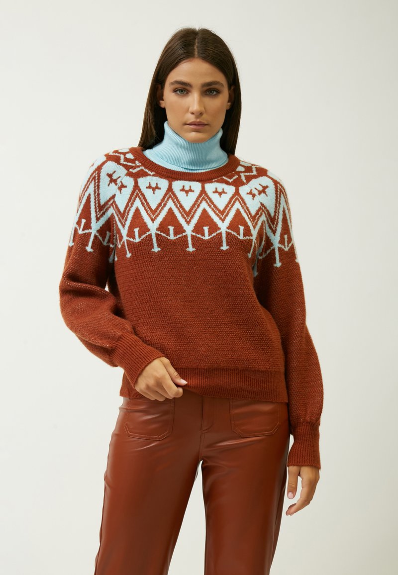 Rustfarbener Strickpullover mit hellblauem Rollkragen, der geometrische Muster aufweist und eine lässige Passform hat, kombiniert mit braunen Lederhosen.