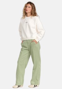 Lichtbeige sweatshirt met geborduurd logo, gecombineerd met groene corduroy broek met zakken en een relaxte pasvorm. Casual schoenen maken de look af.