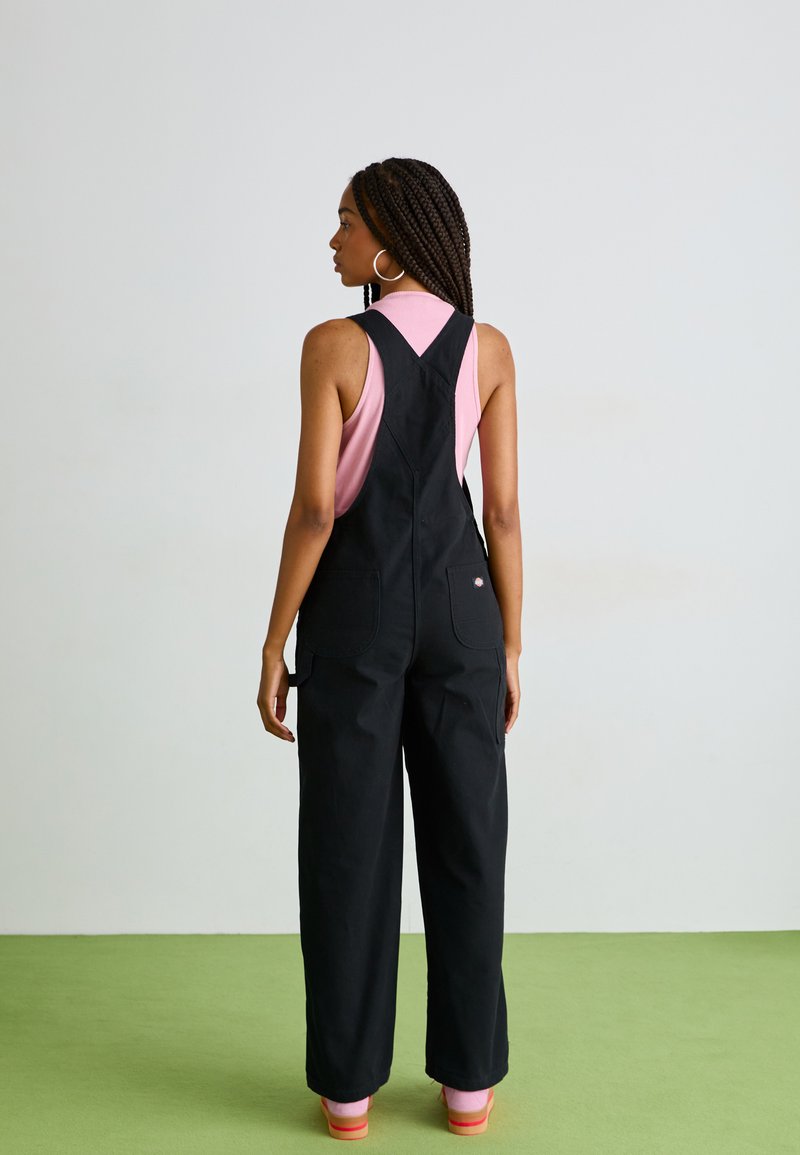 Tuta in cotone nero con gambe larghe, dotata di tasche utili e spalline regolabili, indossata sopra un top senza maniche rosa. Calzature di colore brillante e uniforme.