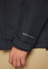 Veste noire imperméable avec le logo OMNI-TECH sur le poignet. Présente une sangle en velcro et un tissu texturé. Poignet doublé de matériel doux.