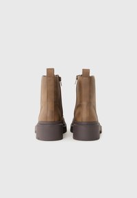 Une paire de bottines en cuir marron avec des fermetures éclair sur le côté et de grosses semelles marron foncé, vues de l'arrière sur un fond blanc.