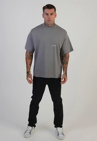 Graues T-Shirt mit kurzen Ärmeln und Rundhalsausschnitt, mit einem kleinen weißen Logo auf der Brust. Kombiniert mit schwarzen Jeans und weißen Turnschuhen.