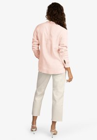 Chemise en lin rose clair avec un col rond et des poignets à bouton, présentant un détail de pli le long du dos. Associée à un pantalon blanc cropped.